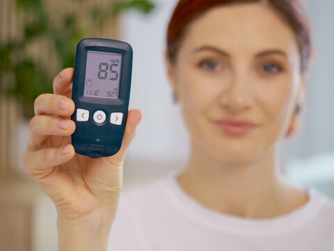 A woman holds a glucometer showing a reading of 85 mg dL. She shares her experience with managing blood sugar and diabetes at her home during the afternoon.