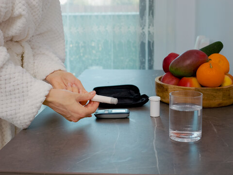 A woman in a kitchen checks her blood sugar level with a glucometer. She prepares to take a blood sample while managing diabetes and monitoring health.