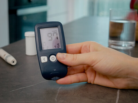 A woman checks her blood glucose meter log with past blood sugar results to monitor her diabetes management in the kitchen.