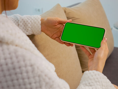 A woman sits on a couch at home and holds a smartphone with a green screen. She looks at the phone while relaxing in her comfortable space.