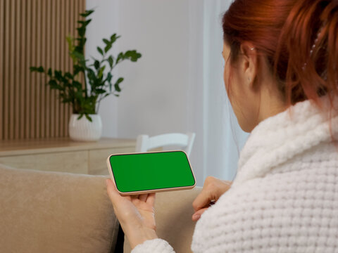 A woman sits on a sofa in her living room holding a phone with a green screen. She focuses her attention on the phone while relaxed at home.