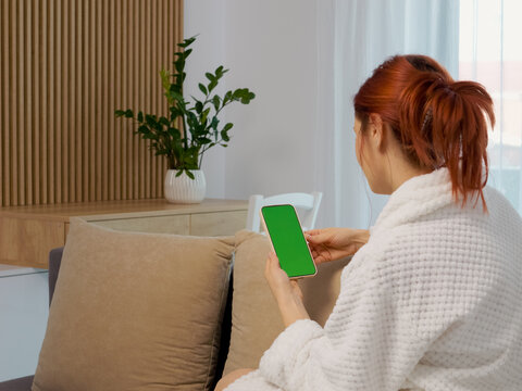 A woman sits on a sofa in her home and looks at a green screen phone. She interacts with it while relaxing in the living room during the day.