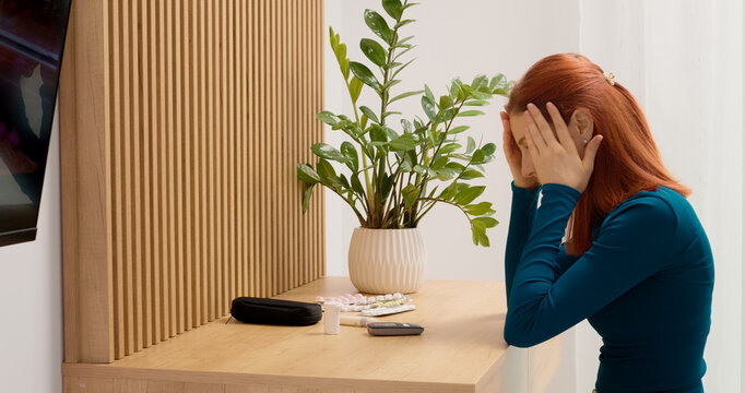 A woman uses a glucometer to check her blood sugar level. She reacts to the result and looks through her medication on the table for options to manage her health.