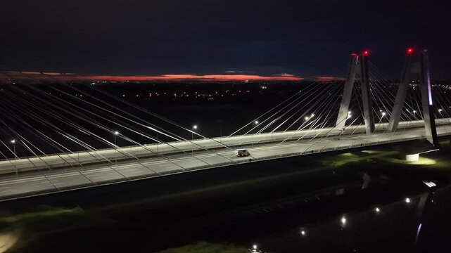 Vehicles crossing over modern cable-stayed bridge at night illuminated by streetlights. Cars riding on a dark road. Autos drive at evening highway. Aerial view