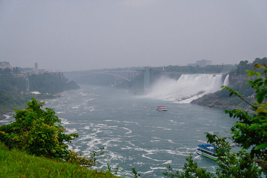View of Niagara Falls with the Rainbow Bridge in the background, mist rising from the cascading water, and tour boats navigating the river in Niagara Falls, Ontario, Canada.