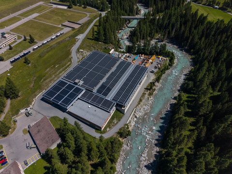 Aerial view of the Upper Engadine Wastewater Treatment Plant featuring extensive rooftop solar panels, situated beside the turquoise waters of the Inn River in S-chanf, Grisons, Switzerland.
