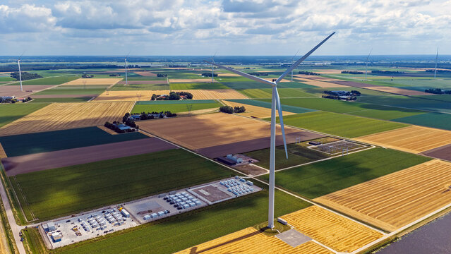 Aerial view of a large wind turbine standing amidst a patchwork of golden and green agricultural fields with power infrastructure in Dronten, Flevoland, Netherlands.