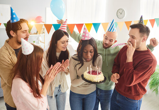 Happy birthday cake, multiethnic friends in funny caps, company congratulate black woman coworker with birthday, friendly presenting gift to happy girl on home party, funny celebration to amuse 