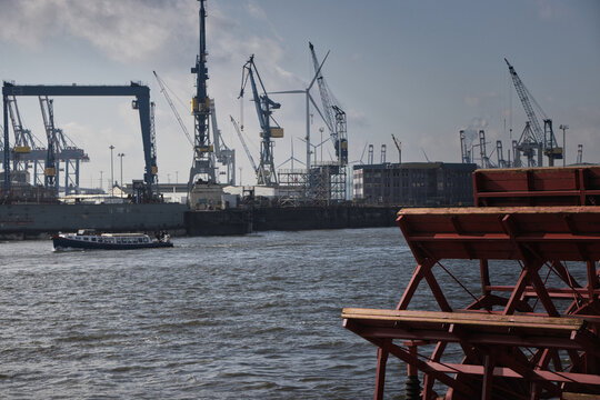 Blick &uuml;ber ein Gew&auml;sser auf den Hamburger Hafen mit mehreren Verladekr&auml;nen und Schiffbauanlagen. Im Vordergrund rechts befinden sich rostrote Holzgestell eines Raddampfers