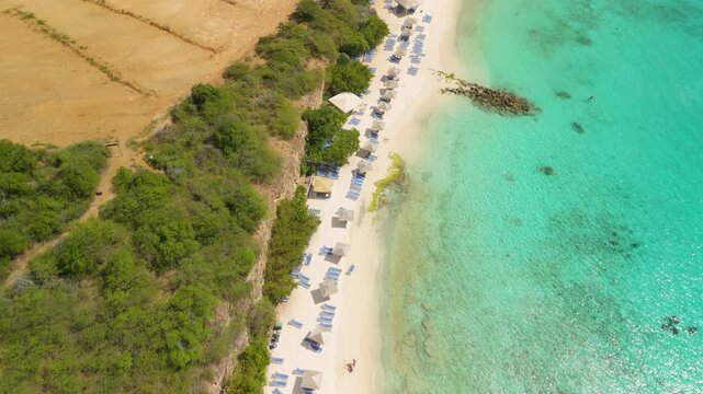 Rows of blue loungers and thatched huts line sandy shore beside wooden pier and solar panels in aerial view of Playa Porto Marie Curacao
