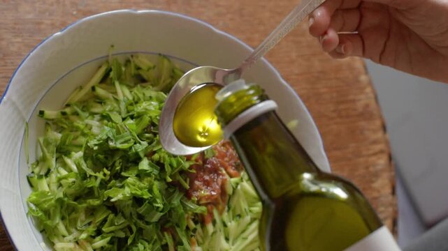 Close oblique top-down shot of right hand holding patterned metal tablespoon while olive oil is poured from tilted green glass bottle, forming glossy pool in spoon above bowl of shredded lettuce and