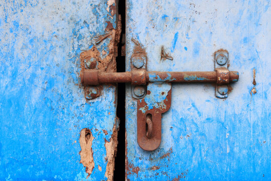 Close-up of a weathered red metal door secured with a padlock, featuring peeling paint and a rustic textured surface