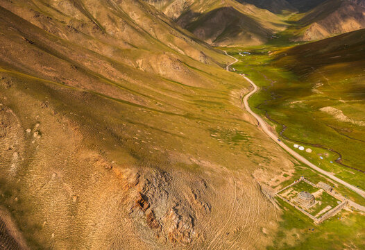 Aerial view of Tash Rabat Caravan Palace nestled in a mountain valley with a winding dirt road, green pastures, and rugged slopes in At-Bashi District, Naryn Region, Kyrgyzstan.