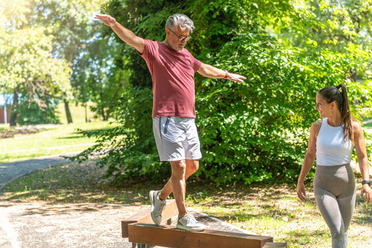 Senior man practicing balance beam walking under the supervision of a fitness coach to improve stability, coordination, body awareness, and functional balance during outdoor training.