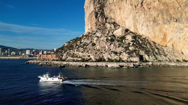 Aerial view of a fishing boat sailing past the iconic Penon de Ifach rock formation with the coastal city skyline in the background in Calp, Valencian Community, Spain.