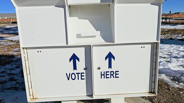 Secure election ballot box with Vote Here arrows, mail-in voting slots, surveillance warning, and Voting Closed sign standing outdoors beside snow-covered ground in bright winter daylight.