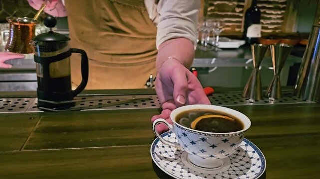 Close-up of a decorative teacup filled with dark tea and citrus garnish on a wooden bar, with bartender hand, French press, glassware, and warm cocktail lounge atmosphere.