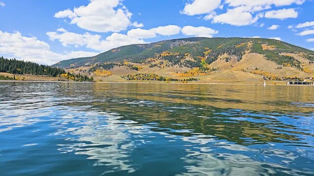 Travel footage from a boat cruising across Dillon Reservoir, Colorado, with glassy blue water, rugged mountain backdrop, golden foliage, and sunny skies reflected on the lake.