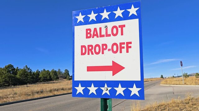 Official ballot drop-off direction sign stands beside a road with bold red lettering, white stars, blue border, and arrow guiding voters during an American election season outdoors.