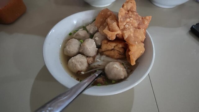 High angle slow motion view of a complete bowl of Indonesian bakso meatballs with fried wontons and spoons placed on a table.
