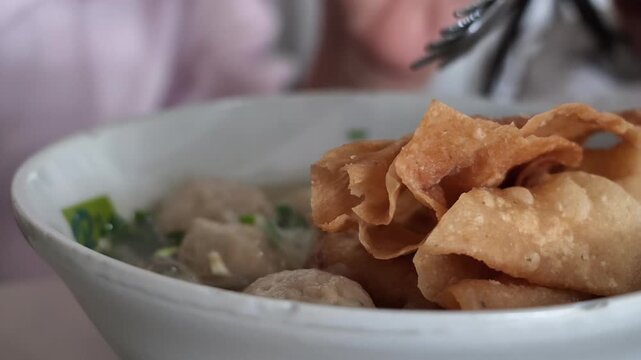 Medium slow motion shot showing a metal spoon scooping clear savory broth from a warm bowl of Indonesian bakso meatball soup.
