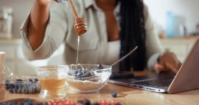 Smiling Black woman adds honey to a bowl while preparing a healthy breakfast in the kitchen and working on her laptop at home.