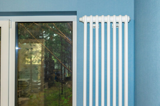 Modern white vertical radiator beside window in blue residential interior. Concept: home heating