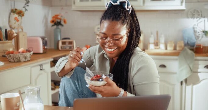 Young Black woman enjoys a healthy breakfast while having a video call on her laptop in the kitchen at home.