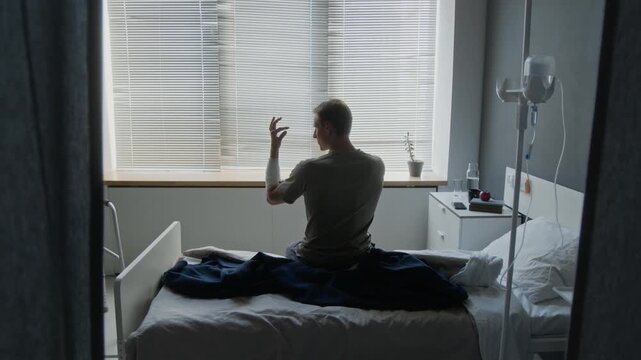 Young adult Caucasian man with bandaged arm sitting on hospital bed, examining his injured hand. Footage for medical insurance, rehabilitation, and patient recovery campaigns