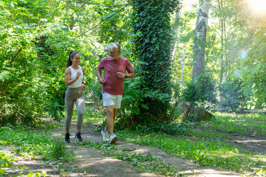 Senior man jogging through a forest park with a fitness coach to improve cardiovascular fitness, endurance, mobility, and support healthy active aging in a natural environment.
