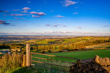 Obraz premium Panoramic golden hour landscape view over the hills and valleys of West Yorkshire, England, with the city of Bradford visible in the distance. A rusty metal farm gate and dry stone wall.