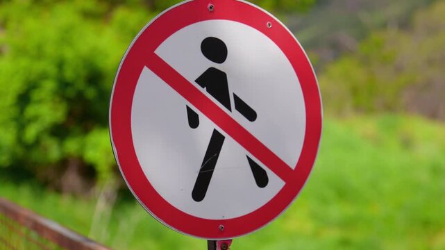 Close-up of a scratched 'no walking' sign with a pictogram of a crossed-out person, indicating a forbidden area for pedestrians against a blurred green background of nature