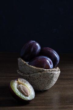 Dark moody still life with fresh purple plums in ceramic bowls and a halved plum with pit