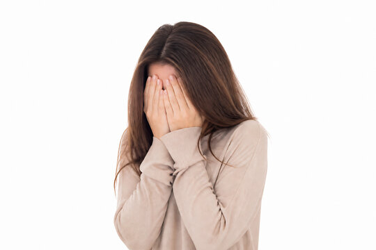 A young woman with long brown hair is covering her face with both hands against a white background.
