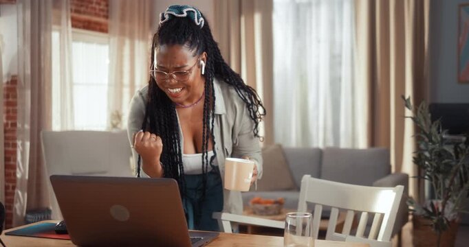 Excited young Black woman reads good news on her laptop and celebrates a personal achievement with a joyful winning reaction at home.