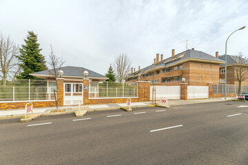 Naklejka premium Suburban brick residential complex with gatehouse, wire fencing, and quiet tree-lined street on cloudy day