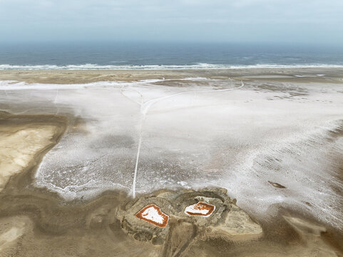 Aerial view of the Skeleton Coast salt pans with distinctive reddish pools and white salt crusts stretching towards the Atlantic Ocean, Dorob National Park, Kunene Region, Namibia.