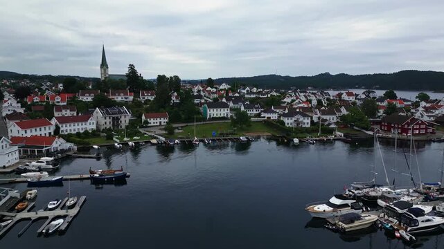 Aerial view of Lillesand harbor with white wooden houses, moored boats, and a church spire under a cloudy sky in Lillesand, Agder, Norway.