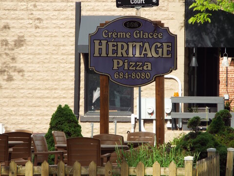 Gatineau,Quebec, Canada &ndash; June 7, 2026: A wooden sign reads "Cr&egrave;me Glac&eacute;e H&Eacute;RITAGE Pizza" outside an establishment featuring outdoor brown patio chairs and green bushes on rue Principale.