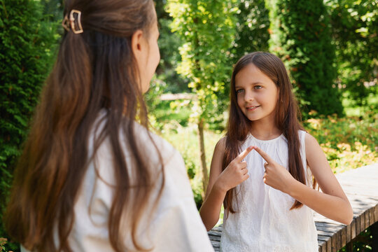 Two children share a moment in a park. One child uses sign language to communicate while the other listens attentively. Sunlight filters through the trees