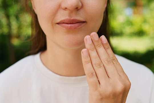 A woman shows a sign language gesture while standing in a park surrounded by green trees. She is focusing on her hand movements in the sunlight