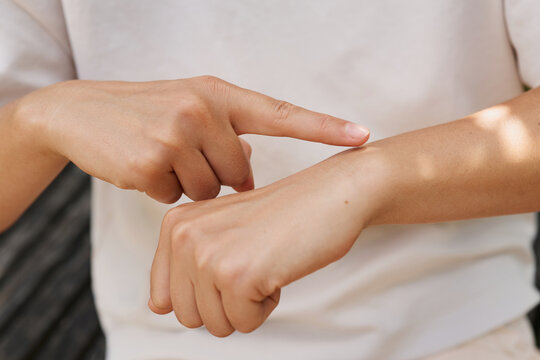 A person is showing a sign language gesture by pointing to their arm while sitting outside. The scene is bright with sunlight filtering through the leaves