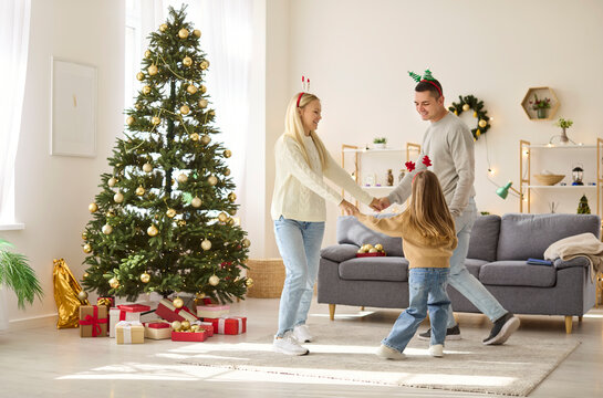 Happy Christmas family dancing near Xmas tree, holding hands to spin together. Parents and child dance to music by gifts, fun home party celebration and winter holidays of father, mother and daughter