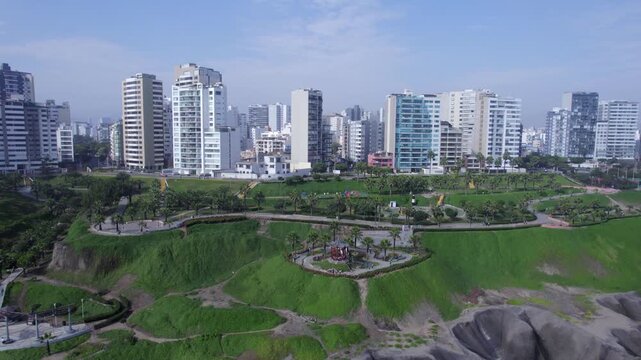 Aerial view of Costa Verde with lush green cliffs, park pathways, and modern high-rise buildings along the coastline in Miraflores, Provincia de Lima, Peru.