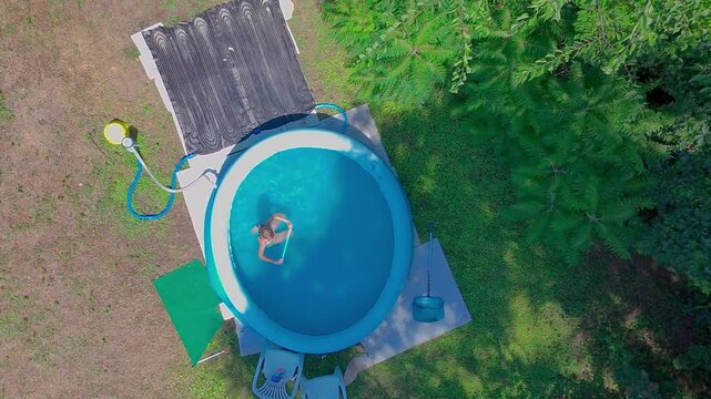 High angle top-down aerial shot captures a pre-teen girl playing and moving joyfully in the clear blue water of a round inflatable swimming pool in a sunny green backyard garden.