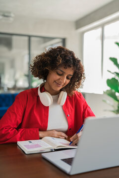Vertical shot of curly haired hispanic woman at notebook, using headphones, writes notes beside laptop. Young indian student stays focused, concentrating on homework study at desk this afternoon.