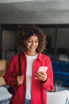 Vertical shot of curly haired hispanic woman carrying backpack, using smartphone, texts with smiling expression. Young latina student checks messages before leaving home this early weekday morning.
