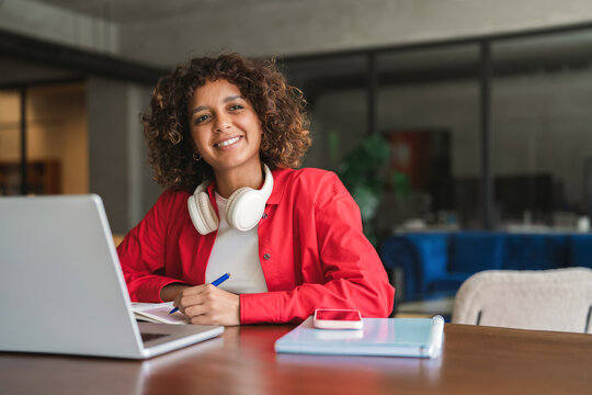 Writing in notebook beside open laptop, curly haired latina woman using headphones, smiling as student at desk. Young hispanic girl looks toward camera during focused study session. Copy space