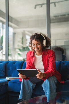 Vertical shot of curly haired latina woman wearing headphones, using tablet, smiling, browsing online course. Young indian student studies from couch near glass table this afternoon. Copy space
