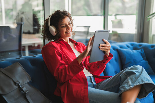 Wearing headphones, curly haired latina woman using tablet, smiling and streaming video with legs crossed on couch. Young hispanic girl relaxes in sunlit loft apartment during quiet afternoon at home.